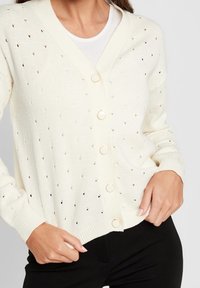 Kvinna iklädd en vit knäppt cardigan med små hål över en vit skjorta och svarta byxor, poserande med händerna nära fickorna.