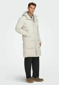 Lichtgrijze gewatteerde jas met een capuchon, voorzien van een knoopsluiting en zijzakken, gecombineerd met donkere broek en bruine schoenen. Glad textuur van de stof.