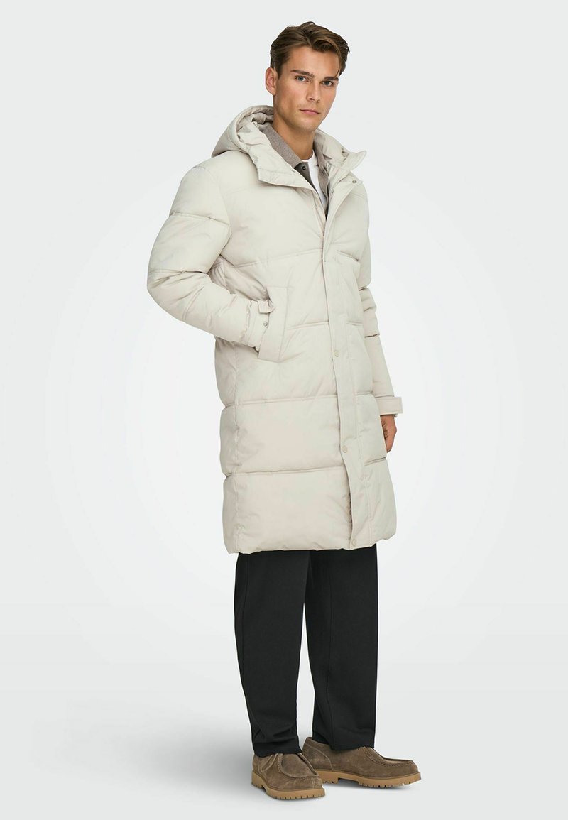 Lichtgrijze gewatteerde jas met een capuchon, voorzien van een knoopsluiting en zijzakken, gecombineerd met donkere broek en bruine schoenen. Glad textuur van de stof.