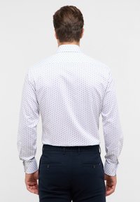 Camicia bianca con un piccolo motivo blu, colletto button-down, maniche lunghe e orlo arrotondato; abbinata a pantaloni scuri blu navy.