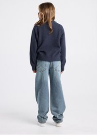 Pull texturé bleu marine à manches longues contrastant avec un jean large bleu clair et des baskets blanches, vu de dos.