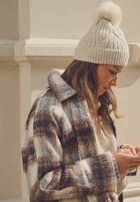Ruudutega villane mantel valgete, roosa ja mustade toonidega; kreemjas kudum beanie pom-pomiga; naine hoiab nutitelefoni.