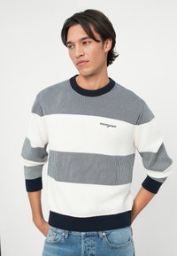 Λευκό και μπλε ριγέ πουλόβερ με ριχτή υφή, διαθέτει στρογγυλή λαιμόκοψη και μανσέτες με ρέλι. Λογότυπο κεντημένο στο στήθος.