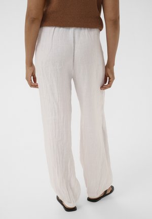 Osoba oblečená v voľných bielych ľanových nohaviciach, hnedom bezrukávovom topu a čiernych sandáloch, stojaca chrbtom k nám na jednoduchom bielom pozadí.