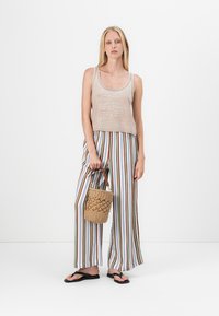 Beige ärmelloses Top, vertikal gestreifte weite Hosen in verschiedenen Farben, hält eine geflochtene Korbtasche, Plastikflip-Flops. Flacher Hintergrund.