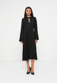 Abito nero di media lunghezza con scollatura a chiave, maniche lunghe e una texture morbida. Abbinato a scarpe con tacchi alti nere.