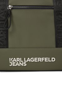 Maslinasto zelena torba od teksturirane tkanine s crnim zatvaračem na zip i istaknutim bijelim logotipom "KARL LAGERFELD JEANS" na prednjoj strani.