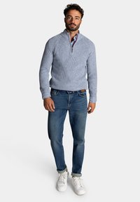 Lichtblauwe gebreide trui met een ritskraag, gecombineerd met donkerblauwe jeans en witte sneakers. De trui heeft een gestructureerd ontwerp.