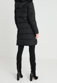 Piumino nero con cappuccio e rifinitura in pelliccia sintetica, presenta sezioni trapuntate orizzontali, abbinato a leggings neri aderenti e stivali alti fino al ginocchio.