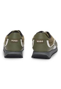 Pár olivově zelených tenisek HUGO s černými podrážkami, bílými akcenty a žebrovanými textilními detaily, zobrazené zezadu na bílém pozadí.