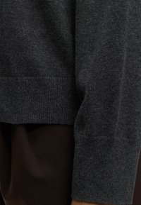 Personne portant un pull en maille gris foncé avec ourlet et manche côtelés sur un pantalon sombre, montrée en gros plan de la taille au poignet.