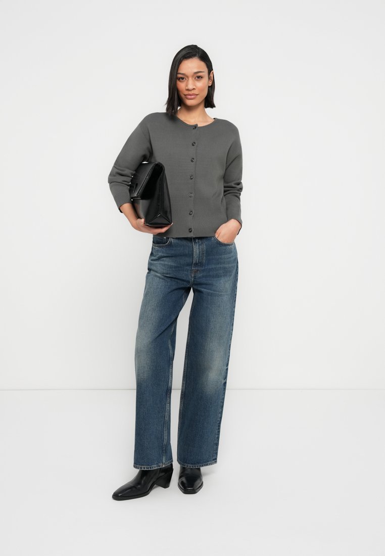 Cardigan gris en maille avec encolure ronde et boutons sur le devant, associé à un jean bleu à jambes larges. Le modèle tient un sac à main rectangulaire noir.