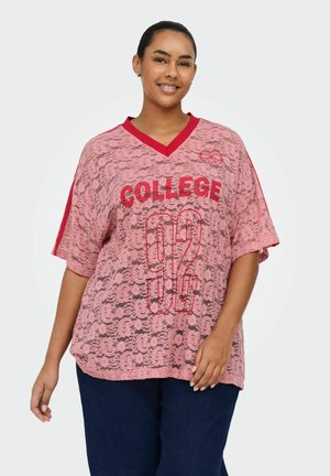 Lächelle Frau trägt pinkfarbenes, übergroßes Spitzenoberteil mit rotem "COLLEGE 92"-Schriftzug, dunkle Hose, Haare zurückgebunden, steht vor einfarbigem Hintergrund.