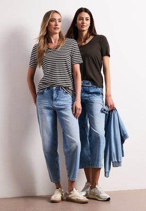 Deux femmes portant des jeans bleus décontractés et des t-shirts se tiennent côte à côte contre un mur blanc ; l'une tient une veste bleue.