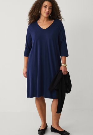 Kvinde med krøllet hår iført en marineblå V-hals kjole, sorte flade sko, guldbøjle, og holder en sort sweater i højre hånd.