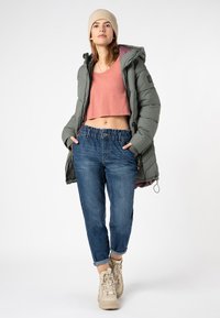 Jonge vrouw in een beige beanie, groene gewatteerde jas, roze crop top, blauwe spijkerbroek en beige veterschoenen, staand met haar handen in haar zakken.