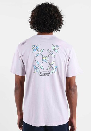 T-shirt à manches courtes lavande clair avec un motif floral bleu et vert au dos, accompagné du texte "OXBOW" en dessous. Matière en coton doux.