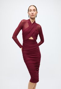 Robe bordeaux avec manches longues en mesh, texture froncée et détail découpé. Le tissu semble extensible et ajusté, mettant en valeur la silhouette.