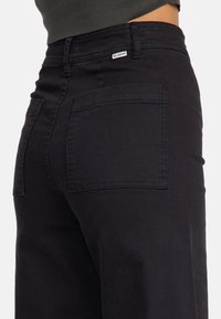 Black denim pants with a high waist, featuring two back pockets, minimal stitching, and a small label detailing the brand.