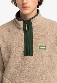Fleece pullover in beige with a green zipper and collar. Features a textured surface, quarter zip, and a logo patch on the left chest.
