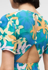 Vue arrière d'une personne portant une robe à fleurs à manches courtes aux couleurs bleu, vert, orange et crème, avec une découpe triangulaire fermée par des boutons en tissu.