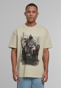 Béžové bavlněné tričko s grafikou postavy v maskáčovém vzoru a dvou psů. Překrývající se prvky zahrnují text ve spodní části a postavu v kapuci.