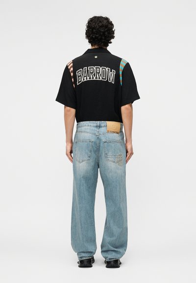 Άνδρας με σγουρά μαλλιά, φορούσε μαύρο μπλουζάκι με κοντά μανίκια με το κείμενο «BARROW» στην πλάτη και ανοιχτό μπλε άνετο τζιν με λεπτομέρεια επιδιορθωτικών πάνελ.