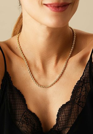 Nahaufnahme einer Frau, die eine gedrehte Goldkettenhalskette und ein schwarzes, mit Spitze verziertes Camisole trägt und sanft lächelt.