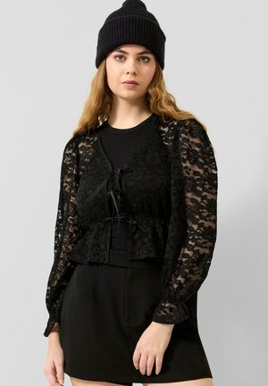 Jeune femme aux longs cheveux ondulés porte un bonnet en tricot noir, un top en dentelle noire transparente sur un t-shirt noir, et une jupe noire, debout devant un fond gris uni.