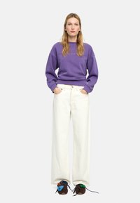 Fioletowa bluza z prążkowanymi mankietami, zestawiona z białymi, szerokimi spodniami typu jeans. Brązowe buty mają kolorowe tkaninowe akcenty na sznurowadłach.