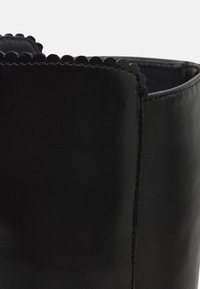 Bottine noire en cuir avec un détail en bord festonné. Texture lisse et forme structurée accentuées par des coutures précises.