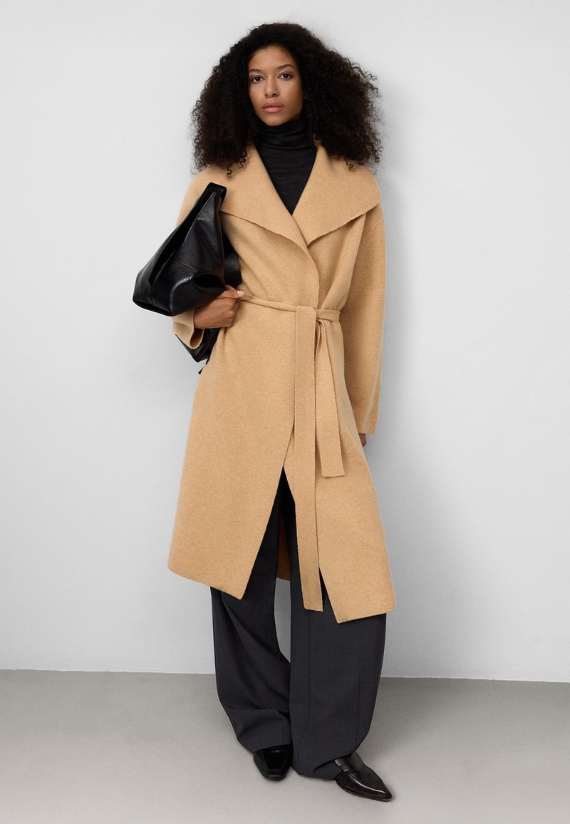 Cappotto avvolgente beige con ampio colletto e cintura, realizzato in tessuto morbido. La modella tiene in mano una borsa nera, indossando un dolcevita nero e pantaloni grigi.