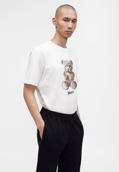 Homme aux cheveux courts portant un t-shirt blanc avec un motif coloré d'ours en peluche et un pantalon noir, debout les mains dans les poches.
