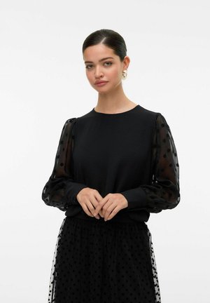 Haut noir avec un corps uni, des manches bouffantes en organza à pois et un col rond. Associé à une jupe noire avec une texture similaire.