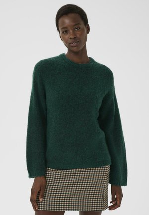 Femme portant un pull en peluche vert foncé et une jupe à carreaux marron, se tenant devant un fond clair uniforme.