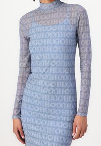 Robe ajustée à manches longues en tissu transparent, arborant un dégradé de nuances de bleu avec le logo "HUGO" répété et des accents de smiley.