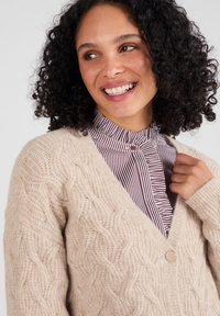 Donna con capelli ricci che sorride, indossando un cardigan lavorato a maglia beige sopra una blusa a righe con colletto alto, mentre tiene il bordo del cardigan.