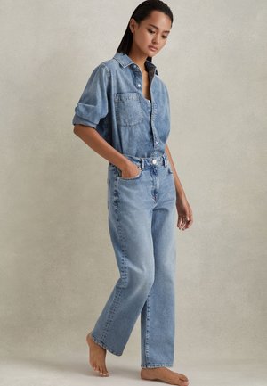 Vrouw staat op blote voeten, draagt een lichtblauw denimshirt met opgerolde mouwen, dat in hoge rechte lichtblauwe spijkerbroek is gestopt.