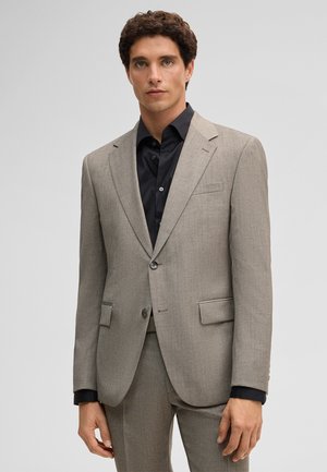 Mann mit lockigem Haar, der eine hellgraue Anzugjacke und Hose über einem schwarzen Hemd trägt und vor einem einfarbigen hellen Hintergrund steht.