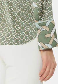 Blusa verde con estampado geométrico, detalles en los puños abotonados y una textura de tela suave. Combinada con pantalones blancos.