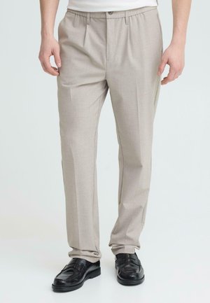 Mann trägt beige plissierte Hose und schwarze Leder-Loafer, steht vor einem schlichten, hellen Hintergrund.