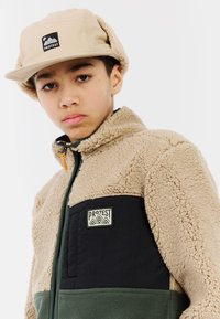 Tan en groene fleecejas met zwarte accenten, voorzien van een rits aan de voorkant, hoge kraag en een logo patch op de borst. Een tan pet complementeert de outfit.