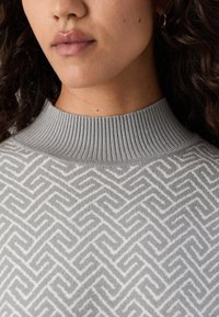 Pull en tricot gris avec un col haut côtelé, présentant un motif géométrique en gris plus clair. Texture douce, design près du corps.