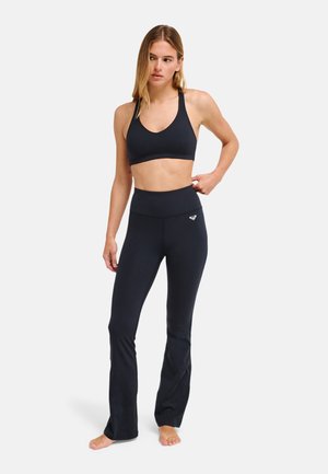 Femme en brassière de sport noire et leggings d'entraînement noirs taille haute, debout pieds nus sur un fond uni.
