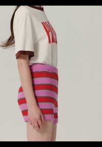 Jeune personne portant un T-shirt blanc avec texte rouge et un short en maille taille haute à rayures horizontales rouges, roses et vertes, debout de profil.