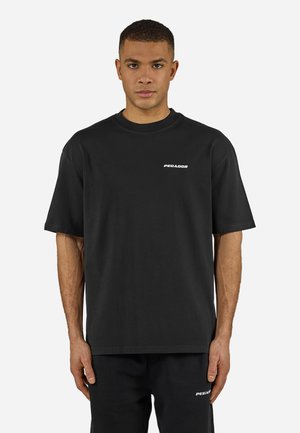 Junger Mann trägt ein schwarzes übergroßes T-Shirt und Hose, beide mit kleinen weißen "PEGADOR"-Logos auf der linken Brust und am Oberschenkel.