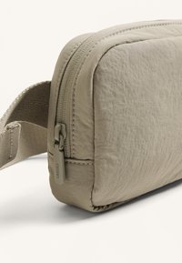 Borsa a tracolla in tessuto beige chiaro con superficie texturizzata, dotata di chiusura a zip e tracolla regolabile per opzioni di trasporto versatile.
