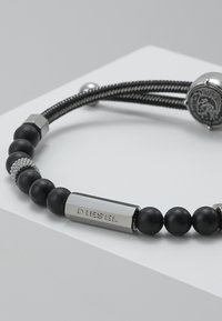 Bracciale nero con perline opache nere e accenti metallici, inclusa una targhetta rettangolare in argento incisa con la scritta "DIESEL". Cordino testurizzato.