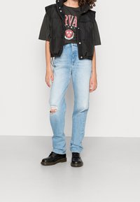 Svart vadderad väst över en mörk grafisk t-shirt, ljusblå slitna jeans med ett hål och svarta snörstövlar.