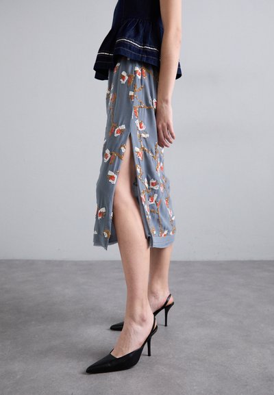 Ανοιχτό μπλε φλοράλ φούστα με πλαϊνές σχισμές, με πορτοκαλί και λευκά κεντημένα διακοσμητικά. Φορεμένη με μαύρες γόβες με μυτερή μύτη.